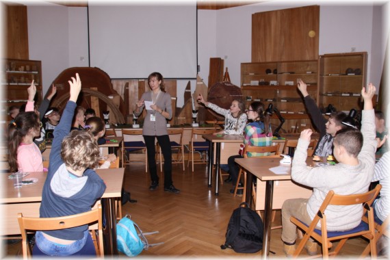 Zajęcia edukacyjne w sali dydaktycznej. Dzieci siedzą w ławkach i podnoszą ręce zgłaszając się do odpowiedzi. W tle stoi kobieta prowadząca zajęcia.